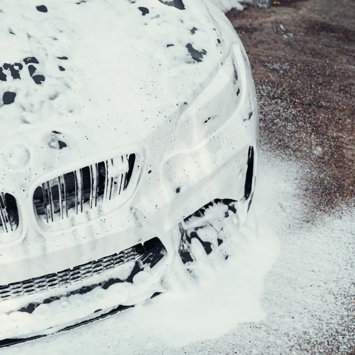 A car, covered in thick, foamy soap, is being washed in an outdoor setting on a concrete ground.
