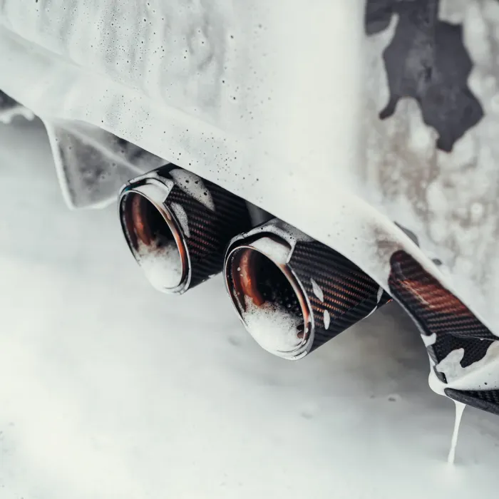 Twin exhaust pipes covered in soapy foam during a car wash, attached to a vehicle with a white foamy exterior.