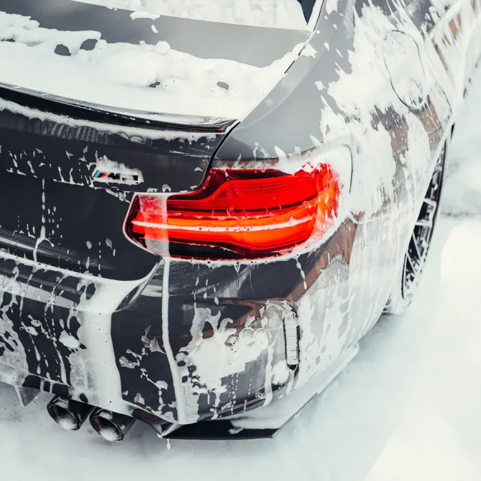 A car's rear corner covered in soap suds, with its brake light illuminated and exhaust pipes visible, during a car wash on a snowy surface.