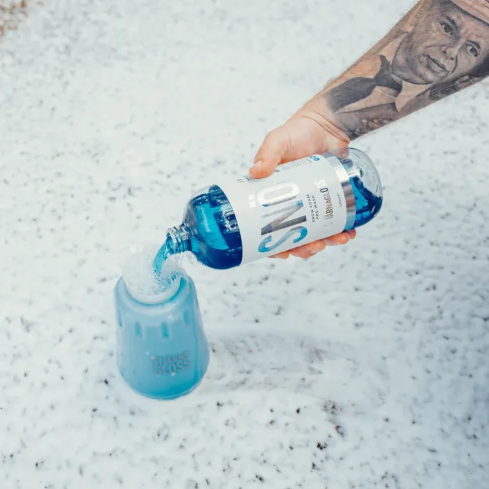 A tattooed arm pours blue liquid from a “SNŌ” bottle into a blue container labeled "Tjarnagloss." The background shows a snowy surface.