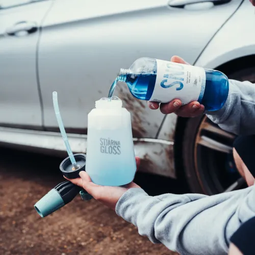 A person pours blue liquid from a bottle labeled "SNO" into a "STJÄRNA GLOSS" container, next to a dirty white car parked on a dirt surface.