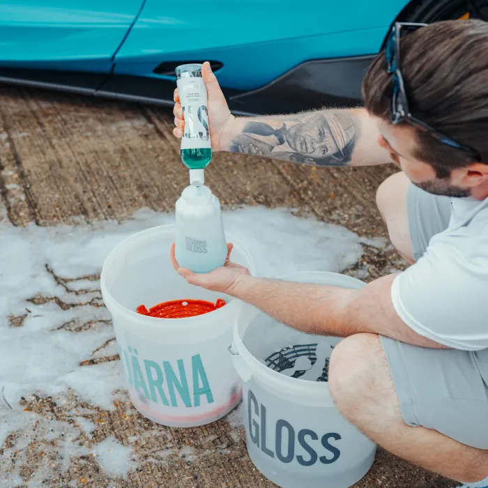 A man pours a cleaning solution into a StjärnaGloss bottle next to two labeled buckets on a soapy, outdoor surface by a blue car.