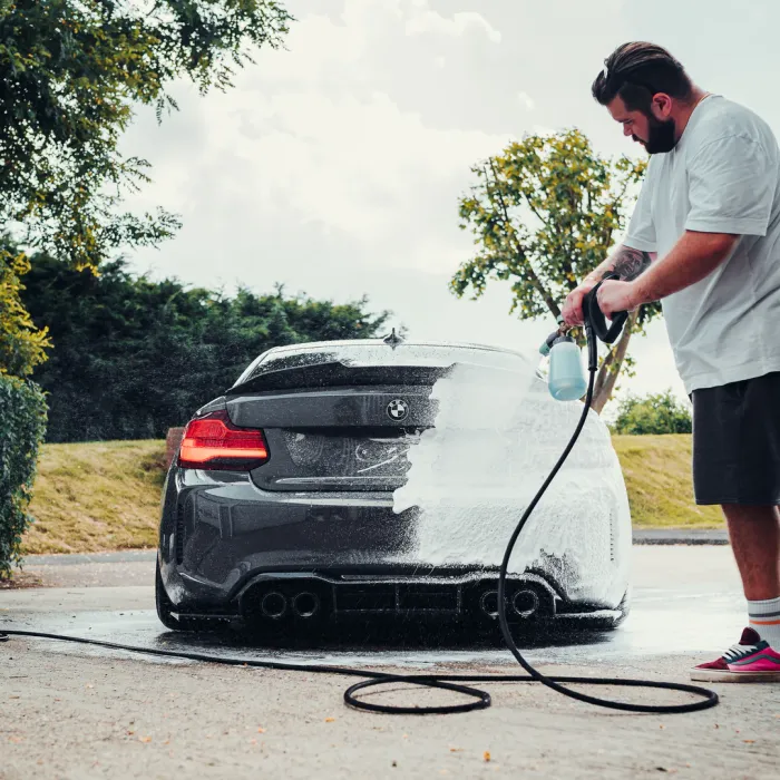 A man sprays foam onto a grey BMW car using a pressure washer, surrounded by lush greenery and a paved ground in an outdoor setting.