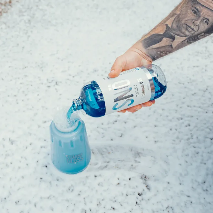 A tattooed arm pours blue liquid from a bottle labeled "SNÖ" into a blue container marked "STJÄRNA GLOSS" on a surface covered in white foam.