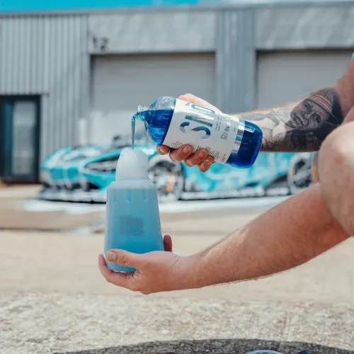 A person pours blue liquid from a bottle labeled "STJARNAGLOSS SNÖ" into a foaming container in a vehicle wash setting with a blue sports car in the background.