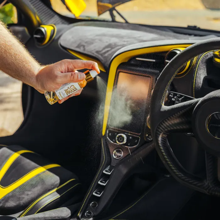 A hand sprays "Stay Fresh" air freshener onto a car's dashboard with black and yellow interior detailing, in a well-lit parking area.