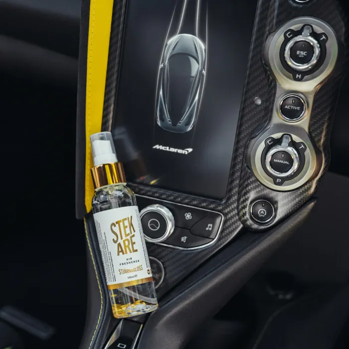 A bottle of "STEK ARE Air Fresheners Stjärnagloss" rests against a car's carbon fiber dashboard console with a touchscreen display showing a McLaren logo and car image.