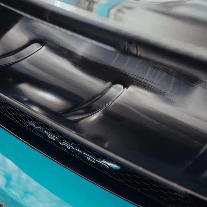 A close-up view of a black car hood with ventilation slits, featuring a logo or emblem with the text "765LT" on a mesh grill, against a blue car body background.