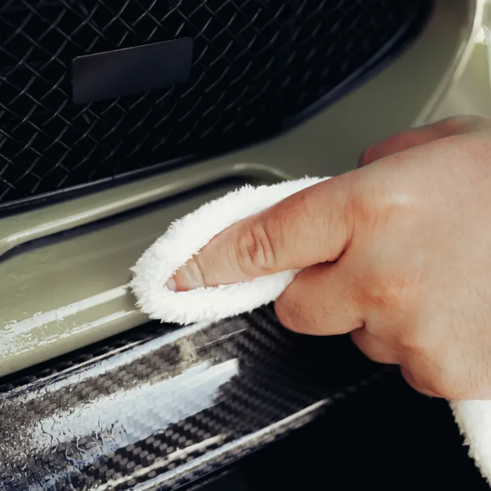 A hand uses a white microfiber cloth to clean the front bumper of a car, with a black mesh grille and shiny, wet surface visible in the background.