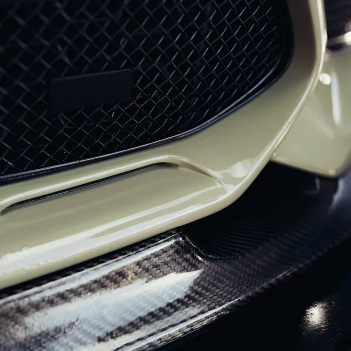 The close-up photograph shows the front grille of a car, with a cream-colored bumper and a carbon fiber lower lip detail, under low lighting conditions.