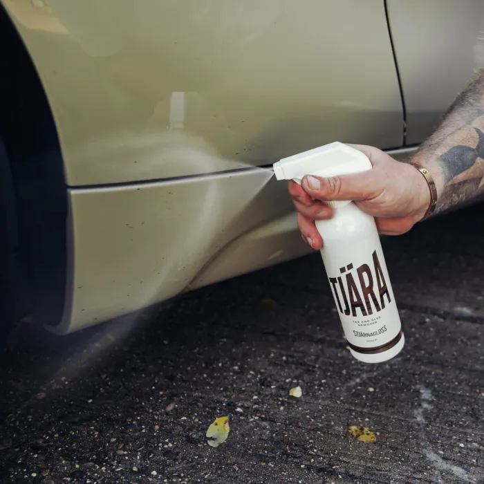 A tattooed hand sprays Tjäragloss Tjära onto a beige car's lower panel, with pavement and small debris scattered around. Text on the spray bottle reads: “TJÄRA, Tar and Glue, Stjärnagloss.”