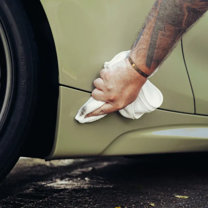 A tattooed arm holding a white cloth wipes a dirty car’s lower panel near the wheel, on a paved surface.