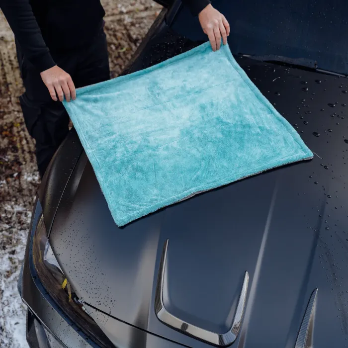 Hands place a blue microfiber cloth on a wet, black car hood outdoors, with foliage and dirt surrounding the area.