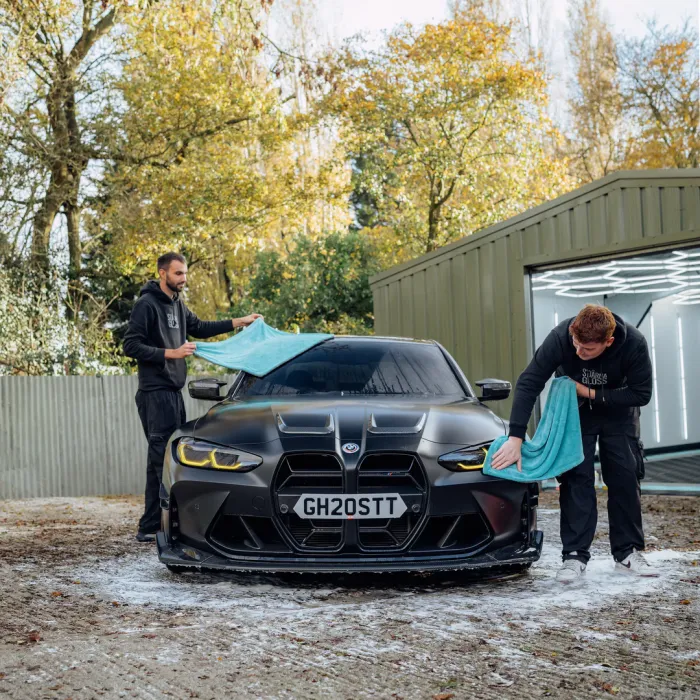 Two men clean a black BMW with teal towels in a driveway beside a green garage. The car's license plate reads "GH20STT". Trees with yellow leaves surround the area.