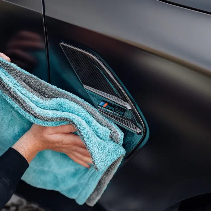 A hand uses a turquoise microfiber cloth to clean a carbon-fiber vent on a black car, bearing the text "M5 competition."