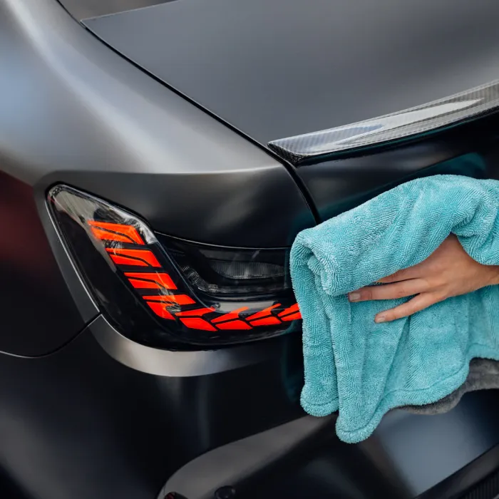 A hand wipes a car's rear light with a turquoise microfiber cloth in a close-up shot, highlighting the car's shiny black finish.