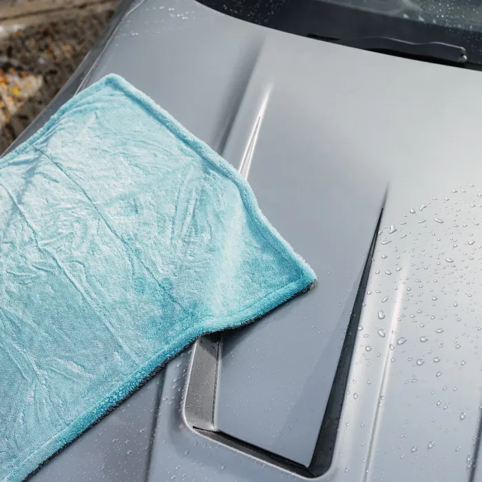 A light blue microfiber cloth lies on the wet, silver hood of a car, showing a cleaning process in progress. The background includes blurred pavement and raindrops.