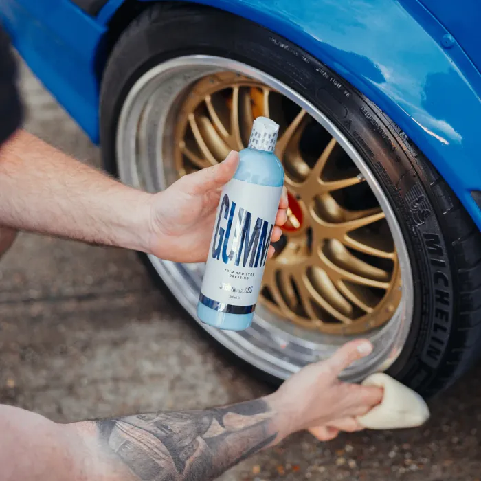 A tattooed individual polishes a car tire with a yellow sponge, holding a bottle labeled "GUMMI - Trim and Tire Dressing - Stjärnagloss" beside a parked blue car on a gravel surface.