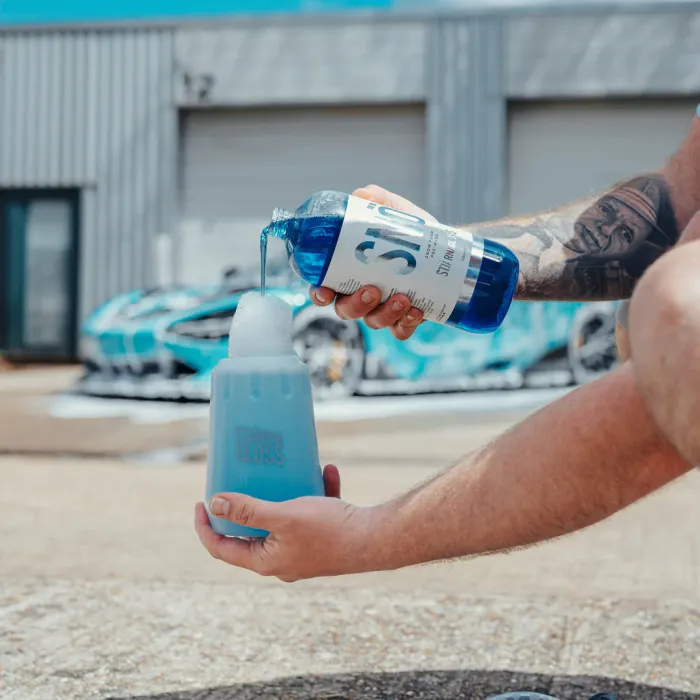 A person pours blue SIO2 wash & gloss liquid into a foaming bottle labeled "TURTLE GLOSS" in an outdoor area, with two sports cars and a grey industrial building in the background.