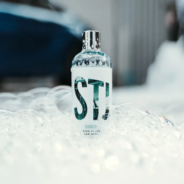 A bottle labeled "STI BUBBLOX HIGH GLOSS CAR WASH" standing amidst foam and bubbles, with a blurred car and grey fence in the background.