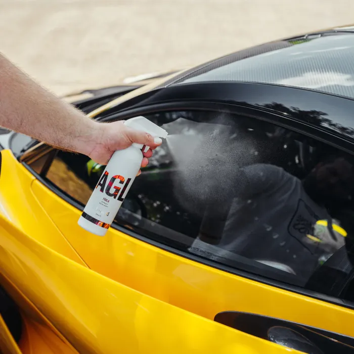 Hand sprays cleaner onto the glass window of a yellow car. The bottle reads "GAGI" and "PEARL". The background is an outdoor setting, likely a parking area.