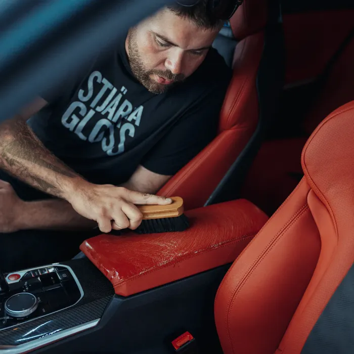 Bearded man wearing "STJÄRNA GLOSS" shirt scrubs a red leather car seat with a brush inside a vehicle.