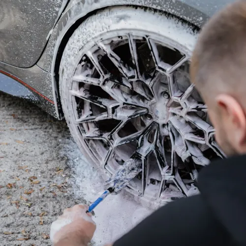 A person scrubs a car's intricate rim with a brush covered in foam, cleaning it outdoors, with the ground partially visible.