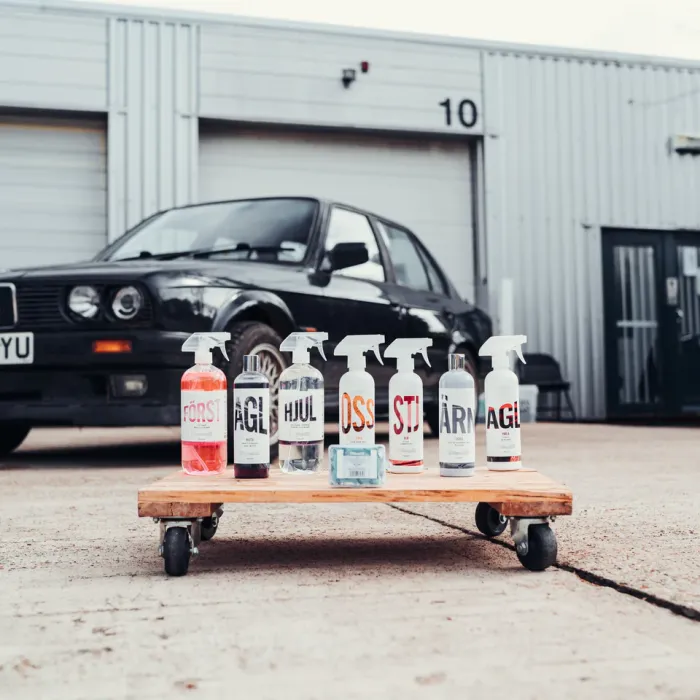Six spray bottles and a bar of soap are arranged on a rolling wooden platform, labeled with "FÖRST," "AGL," "HJUL," "OSS," "STJ," and "ÄR." Behind them, a black car is parked in front of a gray industrial building numbered "10."