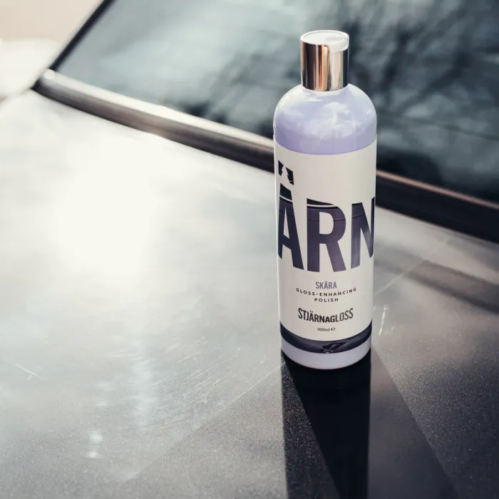 A bottle of Stjärnagloss Skära Gloss-Enhancing Polish sits on a car hood. Sunlight casts reflections, and the car's windshield is visible in the background. Text: "STJÄRNAGLOSS 500ml e."