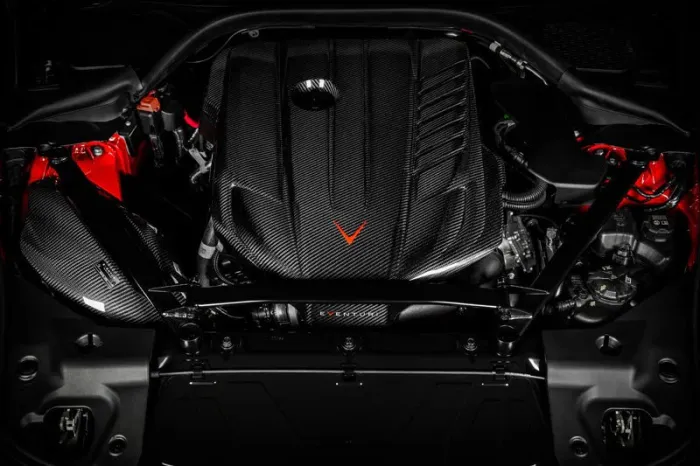 A carbon fiber engine block, adorned with a red V-shaped emblem, is situated within a sleek vehicle hood, highlighting red accents and various mechanical components in the surrounding environment.