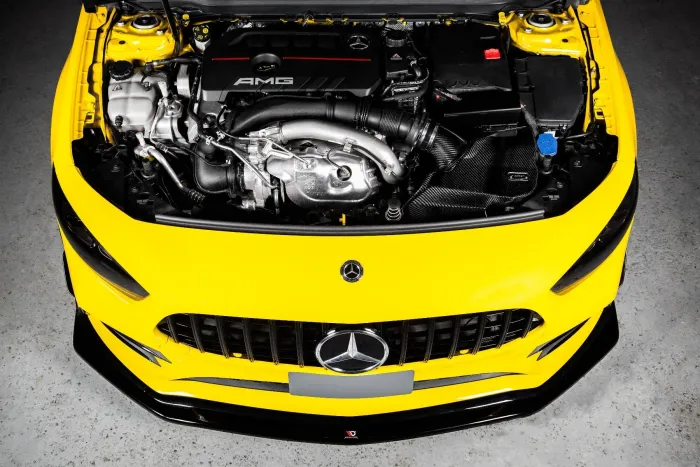 A Mercedes-AMG engine is mounted in a bright yellow car with its hood open, against a concrete floor backdrop. The engine has "AMG" and the Mercedes logo on the engine cover.