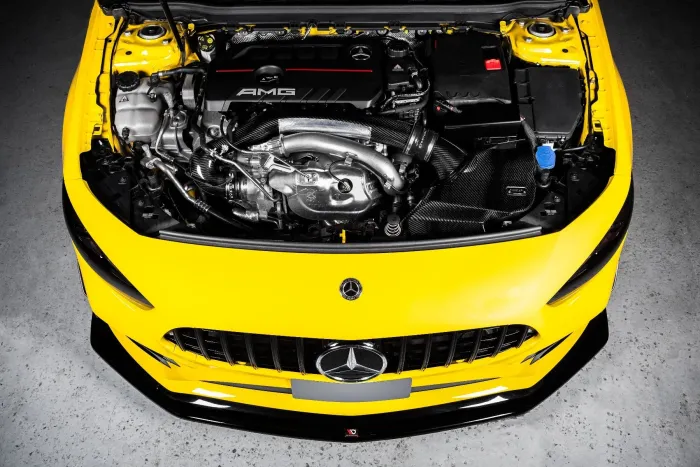 Engine compartment of a yellow car with "AMG" branding on the engine cover, positioned in a well-lit garage setting. The hood is open, revealing various engine components and details.