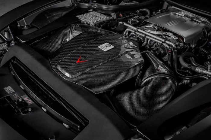 An engine with carbon fiber components situated in a sleek, modern engine bay. The carbon fiber cover features a red arrow and a label with the text "HANDCRAFTED By S. Bucem, Affalterbach."