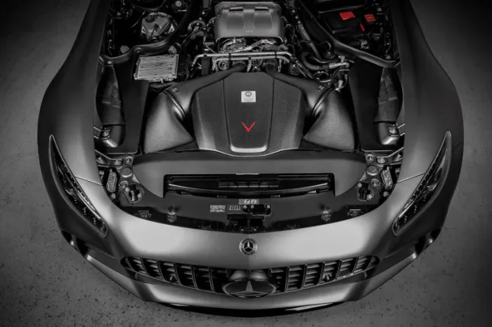 A Mercedes-Benz car with its hood open exposes a detailed engine in a well-lit garage. The engine bay is meticulously organized, showcasing various components, wires, and branding details.