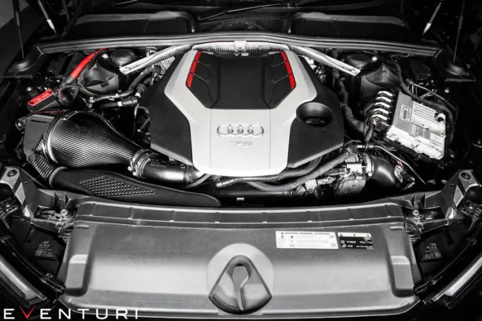 Car engine bay showing an Audi engine with a black and silver cover marked "TFSi," surrounded by various mechanical components, wires, and Eventuri carbon fiber intake. Text: "EVENTURI."
