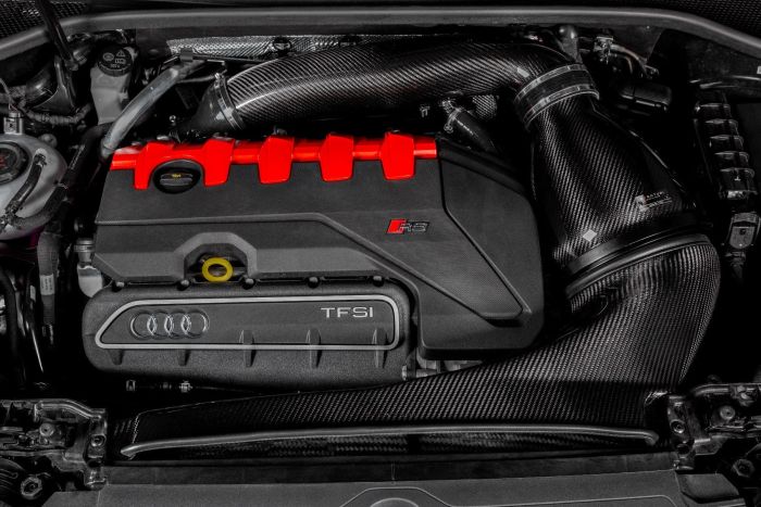 Engine bay with a prominent black and red engine cover showing "RS" and "TFSI" logos, surrounded by carbon fiber air intake pipes and electrical components in a car's compartment.