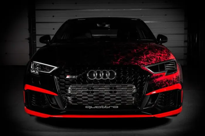 A black Audi car with red accents has a bold front grille displaying the Audi logo and "quattro" text, parked in a dimly lit garage with a closed metallic background.