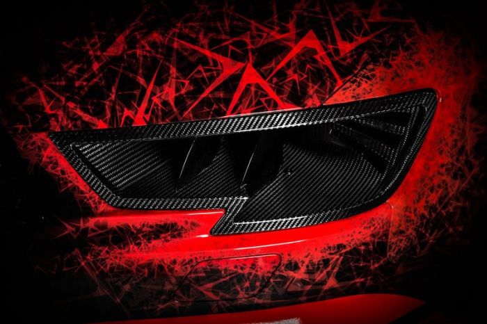 A carbon-fiber car exhaust vent on a red vehicle, surrounded by abstract, jagged red and black patterns, creating a dynamic and aggressive visual effect.