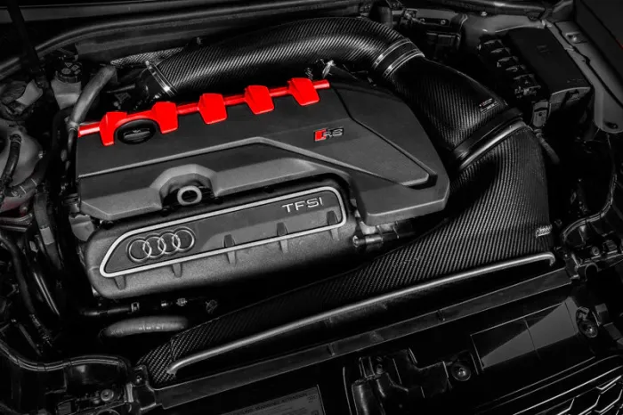 A car engine, with "TFSI" and Audi's four-ring logo on it, features black and carbon fiber components and red accents. The surrounding area includes various engine components and wiring.