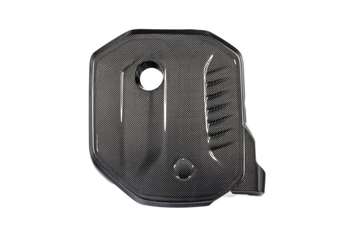 A carbon fiber automotive engine cover with a central circular hole, raised grooves, and sleek patterns, lying against a white background.