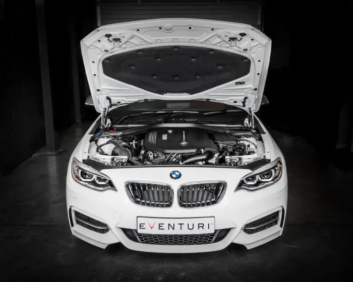 White BMW car sits with its hood open, exposing the engine, in a dimly lit garage. A license plate reads "EVENTURI."