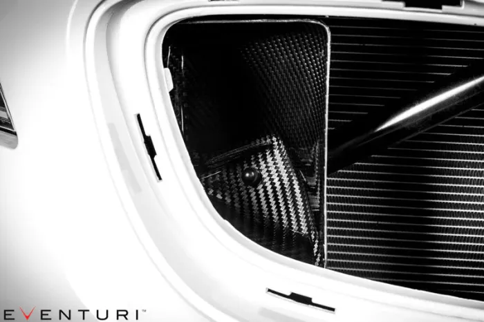Carbon fiber car engine component in a white vehicle's engine bay with branding "Eventuri" in the bottom left corner. Various parts and textures are visible in a tightly framed shot.