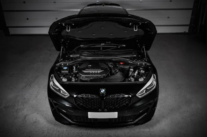 A black BMW car with its hood open, showcasing the engine, is parked in a garage with closed shutter doors in the background.