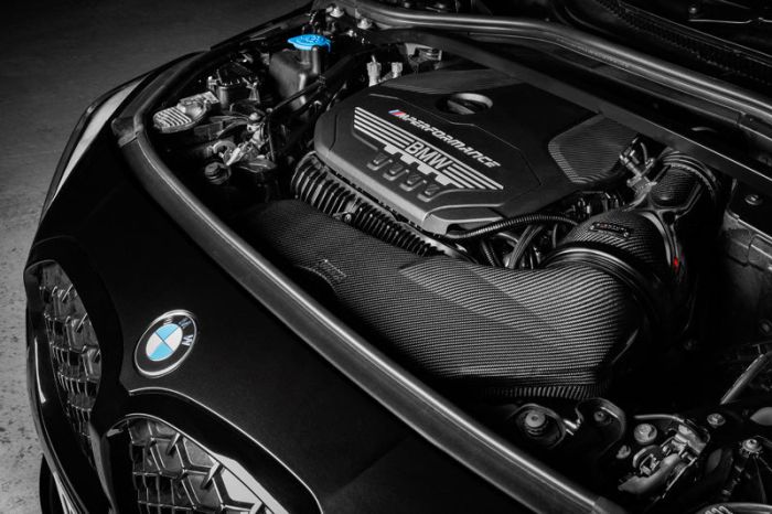 A BMW car engine marked with "M Performance" text is securely fitted in the hood, surrounded by various mechanical components. The car is parked in a dimly lit garage.