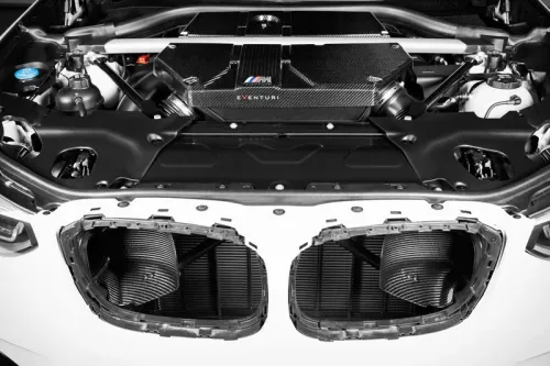 An open car hood reveals a sleek, carbon fiber engine cover labeled “Eventuri” and an M logo, situated in a clean, organized engine bay with visible air intake areas toward the front.