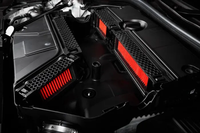 Two air filters with red elements housed in black casing are positioned side by side within the open engine compartment of a vehicle, against a background of various mechanical components.