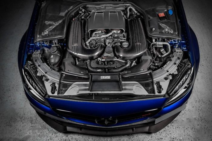 Engine with carbon components positioned within the open hood of a blue vehicle, showcasing various mechanical parts and fine details, placed on a concrete floor in a garage-like setting.