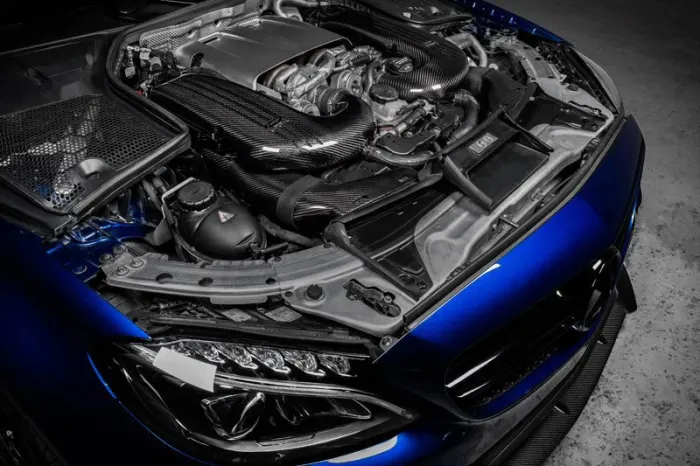 A high-performance car engine with advanced carbon-fiber components is exposed in a blue vehicle parked on a concrete surface.