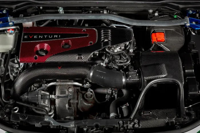 Car engine bay featuring an Eventuri performance air intake system with red and black carbon fiber components, metal braces, hoses, and various mechanical parts, all situated inside the vehicle's engine compartment.
