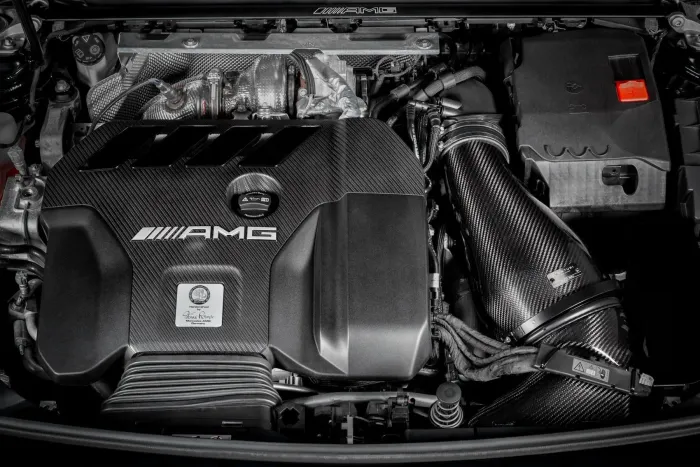 An AMG branded car engine with a carbon-fiber cover, pipes, and various components, displayed within the engine bay of a vehicle. Text: "AMG, Handcrafted by Marco Roger AMG Germany."